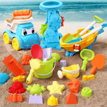 Colorful assortment of plastic beach toys including a toy truck, sand molds, shovels, rakes, and a wheelbarrow on sand near ocean