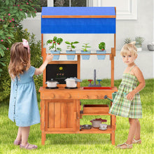 Two young girls playing with a wooden outdoor play kitchen set with pots, a sink, and artificial plants under a blue canopy