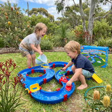 Two young children playing with a blue water park toy set on grass surrounded by green plants and trees