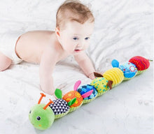 Baby crawling on white bed reaching toward colorful soft caterpillar toy with various textures and patterns